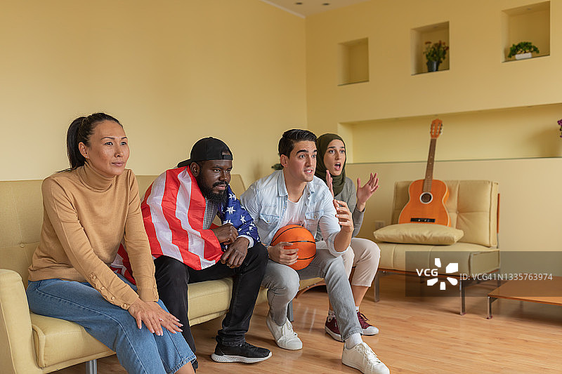 来自美国不同种族的年轻朋友们兴奋地坐在现代公寓里，观看电视上的篮球比赛。图片素材