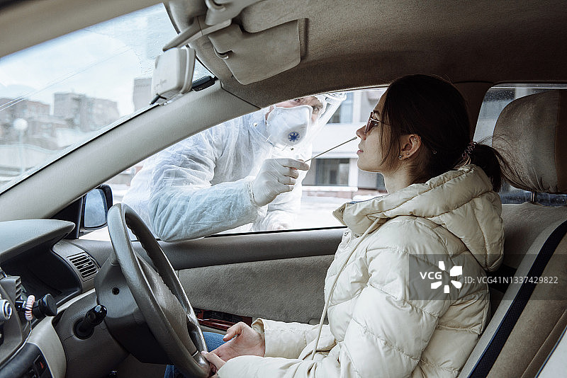 穿着个人防护用品防护服的病毒学家或医生用棉签从一名女司机的车中提取PCR检测样本。新型冠状病毒移动检测概念图片素材