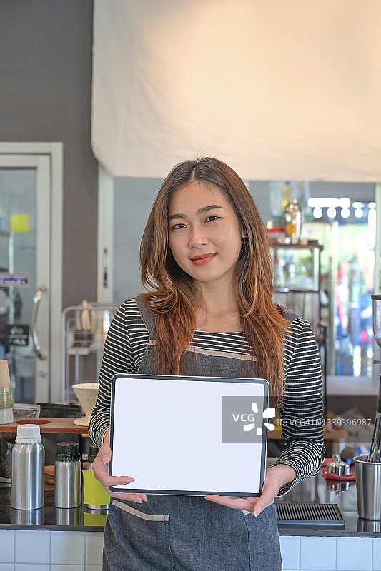 一名快乐的女服务员站在咖啡店里，在空着的屏幕上展示着数码平板电脑。图片素材