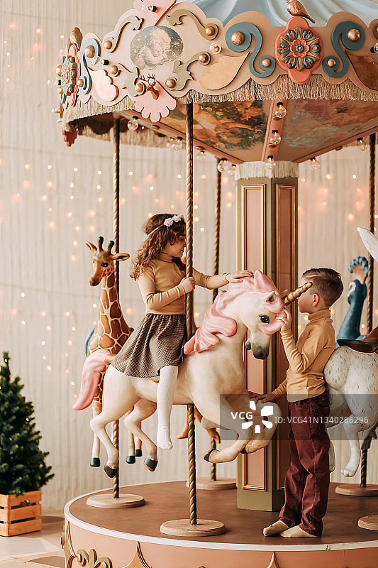 有趣的孩子，十几岁的朋友，一个小女孩和一个小男孩一起骑在旋转木马上，在花环和圣诞树的背景下，在游乐园的新年图片素材