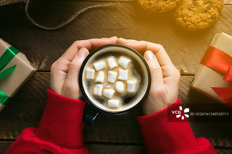 一个穿红毛衣的女孩或女人，手里拿着一杯热咖啡和棉花糖。旁边放着巧克力饼干、圣诞礼物，背景是一张木桌。全家准备过节的概念，平安夜快乐图片素材