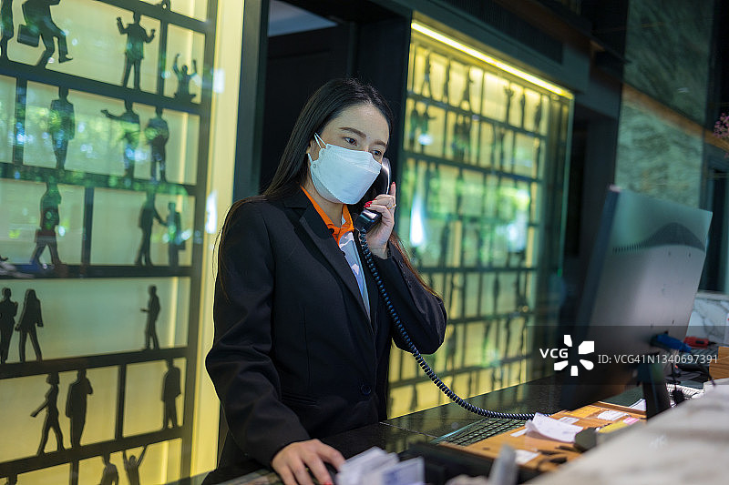 亚洲女酒店员工在酒店前台带着防护口罩工作。图片素材