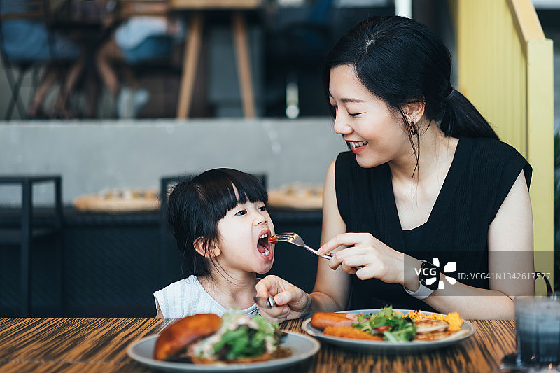 年轻的亚洲家庭在咖啡馆吃午餐。妈妈和她的小女儿分享食物，一起享受一顿快乐的晚餐。家庭和外出就餐的生活方式图片素材