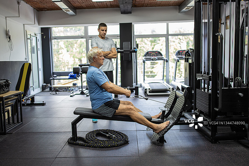 在健身房里，一名年长的男子在健身机上锻炼，旁边是私人教练图片素材