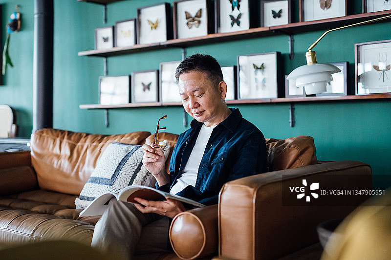 资深亚洲人坐在客厅的沙发上，放松和阅读一本书在家里图片素材