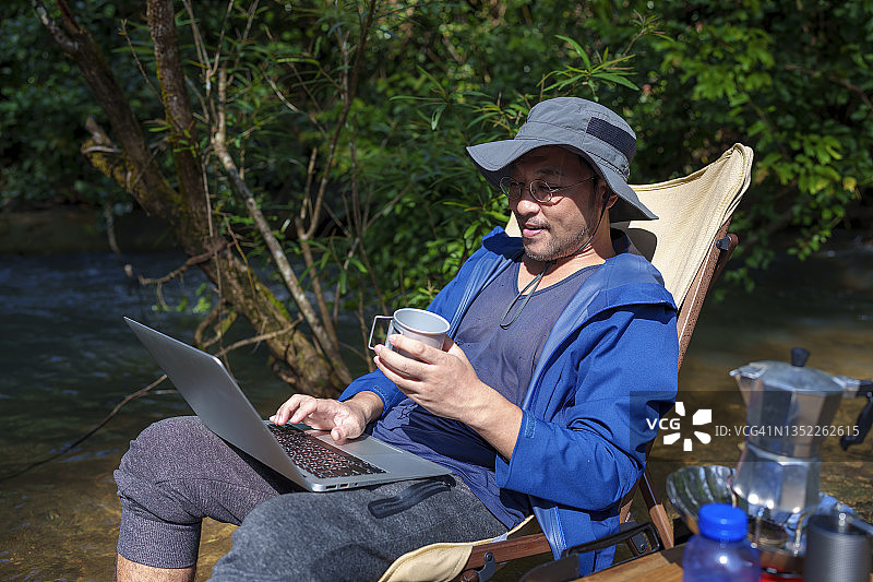 亚洲日本男子拿着咖啡杯坐在河边的露营椅上用笔记本电脑。图片素材