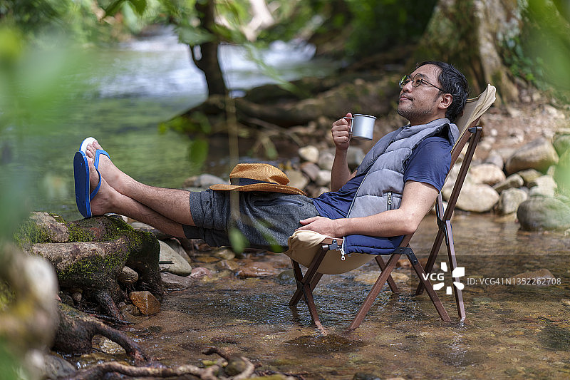 亚洲日本人拿着咖啡杯坐在河边的露营椅上。图片素材