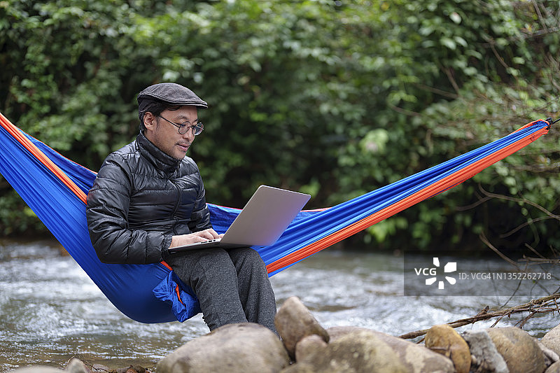 在河边的森林里，一个亚洲日本人坐在吊床上用笔记本电脑。生活方式与现代科技的工作和旅行的概念。图片素材