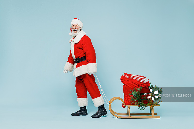 全身快乐的圣诞老人男人50多岁，戴着圣诞帽，红色套装，衣服，带着礼物，雪橇，孤独地行走在素色背景的工作室。2022年新年快乐，庆祝圣诞快乐。图片素材