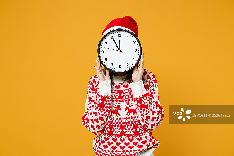 年轻的圣诞老人20多岁，戴着红色的毛衣圣诞帽，双手捂着脸，与时钟隔离在黄色的背景上，工作室肖像。新年庆祝快乐节日概念。图片素材
