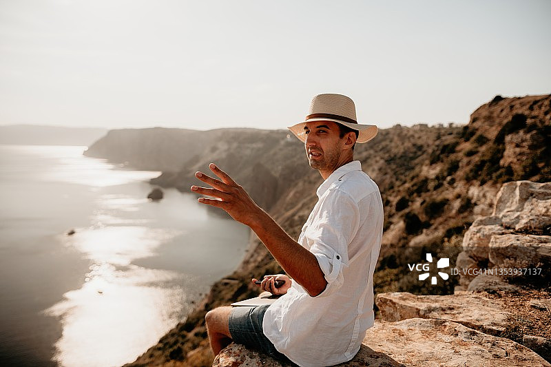 数字游牧民，戴帽子的男人，日落时坐在海边岩石上拿着笔记本电脑的商人，在网上进行交易。假期远程工作图片素材