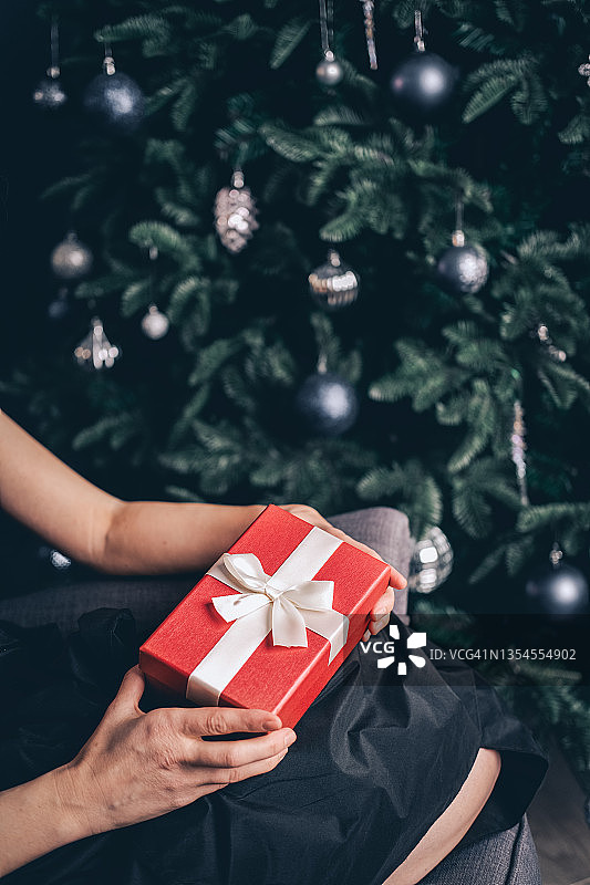 女人手里拿着蝴蝶结的礼品盒。女人的手拿着礼品盒和蝴蝶结靠在圣诞树上。新年庆祝概念。特写镜头。图片素材