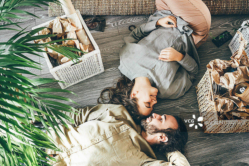 可爱的年轻女人和男人在便服躺在地板上亲吻靠近盆栽棕榈树和箱子里的物品从上面看新公寓。图片素材