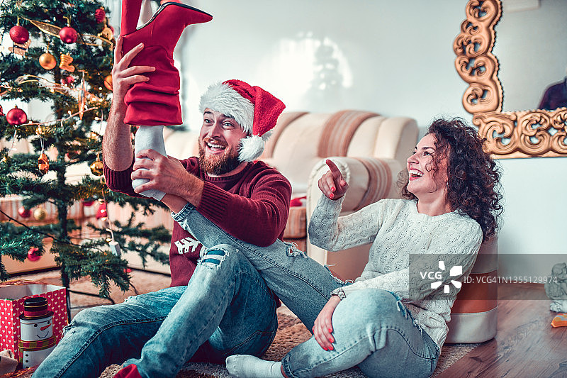 戴着圣诞帽的微笑的男性把圣诞鞋礼物放在树下女朋友的脚上图片素材