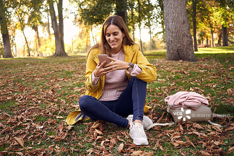 穿着便服和黄大衣的年轻女子在树木环绕的公园里坐着用手机。图片素材