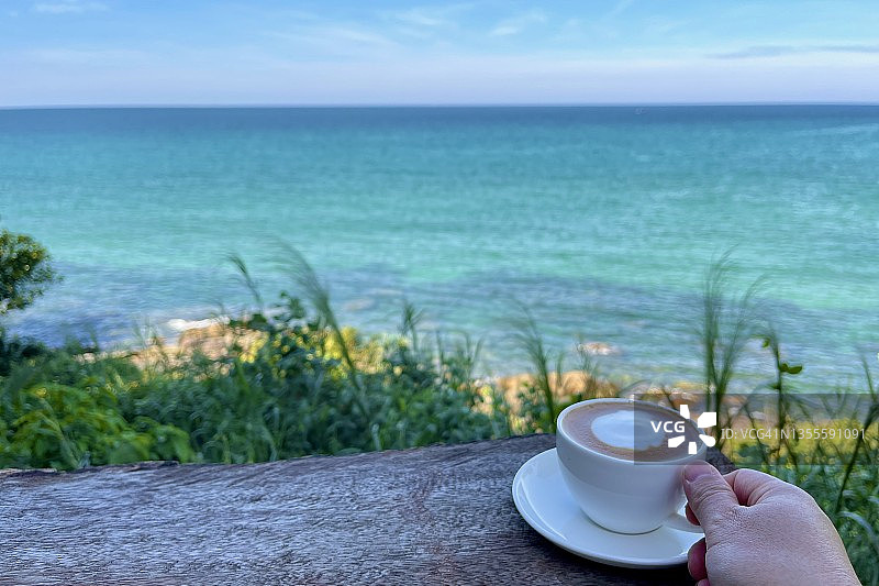 右手拿着一杯咖啡，放在户外的木桌上，以大海和晴朗的天空为背景图片素材