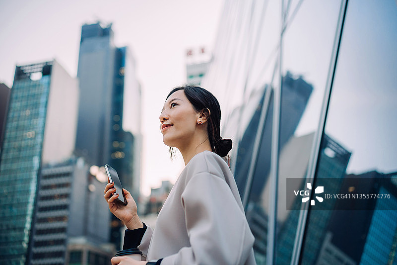 自信而雄心勃勃的年轻亚洲女商人，拿着智能手机看向别处，站在城市金融区的当代企业摩天大楼前。生活方式和技术。女人在工作。女性领导理念图片素材