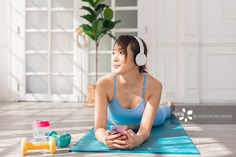选择运动音乐:戴着耳机的亚洲女人坐在运动垫子上，在手机上设置播放列表图片素材