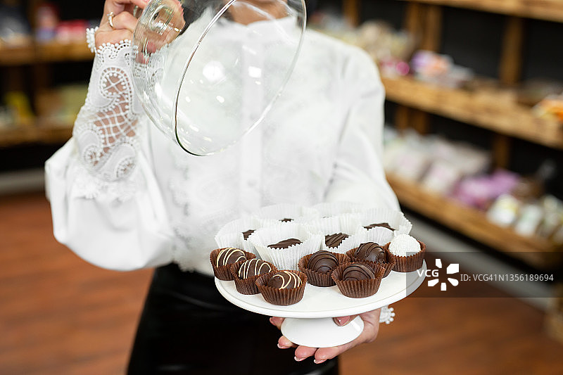 在商店里，一位漂亮的年轻女士的手放在托盘上的玻璃盖子下的手工巧克力。图片素材