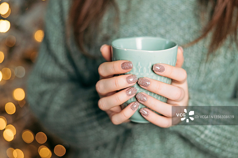 穿着针织羊毛衫的妇女手里拿着一个杯子，里面盛着热巧克力或咖啡。圣诞彩灯亮起，节日气氛温馨。特写镜头图片素材