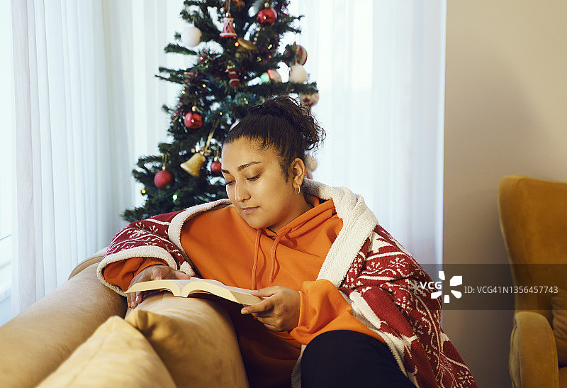 年轻女子拿着圣诞毯子看书，坐在家里的沙发上休息。图片素材