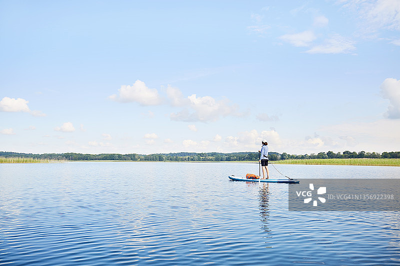 夏天，女人站在田园诗般的湖面上划水，平静的水面上倒映着云朵和蓝天图片素材