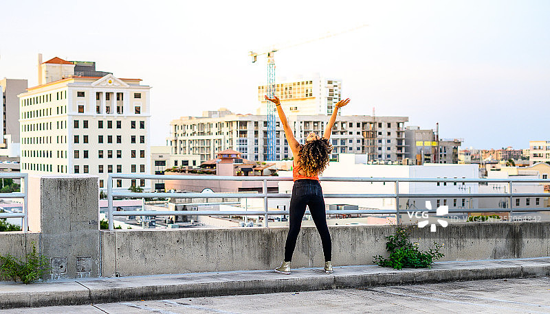快乐的年轻黑人妇女举起她的手臂，俯瞰城市的天空图片素材