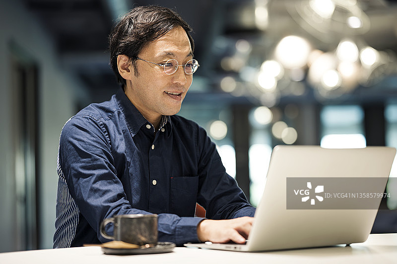 技术驱动业务增长。一个日本商人在办公室用笔记本电脑打字和回复电子邮件给他的客户。图片素材