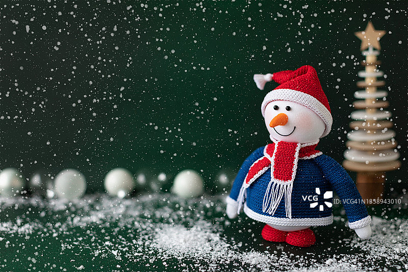 戴着红色帽子和围巾的圣诞雪人站在深绿色背景的雪地里。新年贺卡设计图片素材