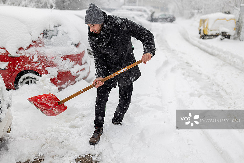 年轻人拿着雪铲。积雪下的汽车和街道。图片素材