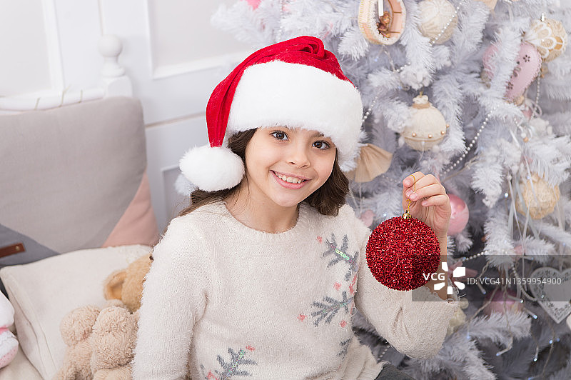 戴着圣诞帽的孩子在装饰圣诞树。华丽的装饰。营造喜庆的氛围。孩子用红球装饰圣诞树。正在装饰圣诞树的小女孩。珍贵的假期活动图片素材