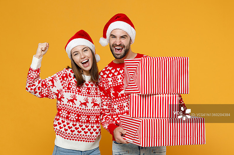 快乐的年轻的圣诞老人夫妇朋友，男人，女人，在毛衣圣诞帽，拿着礼物盒做赢家的姿态孤立在黄色背景。祝新年庆佳节快乐理念。图片素材