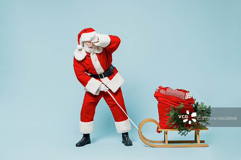 全身疲惫悲伤的老圣诞老人50岁老人戴着圣诞帽红色套装衣服拉着沉重的礼物雪橇出汗孤立在朴素的蓝色背景工作室新年快乐2022年圣诞快乐贺圣诞概念。图片素材