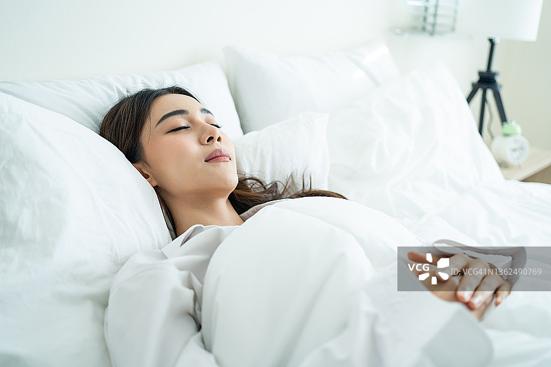 亚洲漂亮舒适的女孩穿着睡衣从卧室里睡觉起来。有吸引力的年轻女子躺在床上感到高兴，舒适的枕头和毯子享受清晨醒来后在房间在家里图片素材