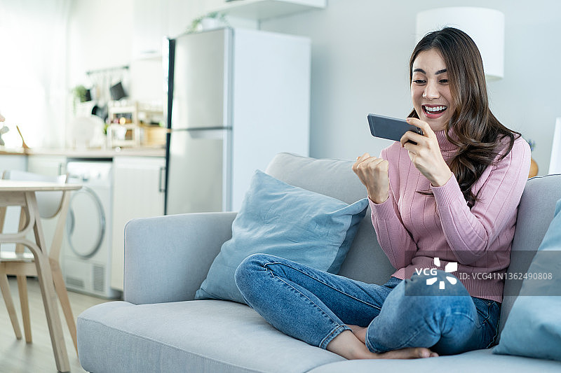 亚洲年轻美女在家玩智能手机游戏。有吸引力的休闲女孩感到快乐和放松，坐在沙发上有乐趣的触摸屏幕上的电话，播放视频享受胜利的房子图片素材