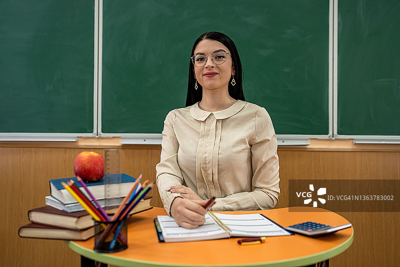 戴眼镜的漂亮老师坐在书桌前，拿着书、铅笔、笔记本、苹果对着黑板图片素材