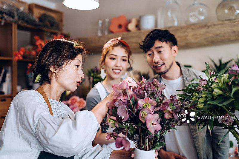 亚裔女性花商，小型花店老板，在花店中协助和与年轻夫妇顾客选购鲜花图片素材