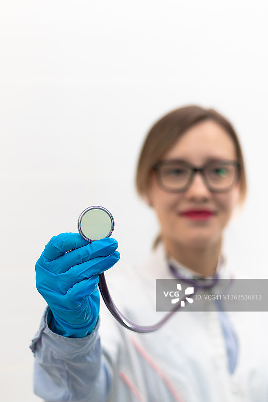 年轻漂亮的女医生，戴着医用手套和眼镜，在医院的白色背景上戴着听诊器。有选择性的重点。肖像。图片素材