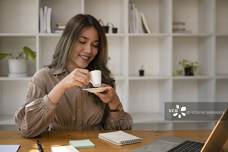 积极的年轻亚洲妇女坐在办公室办公桌和喝咖啡在早上。图片素材