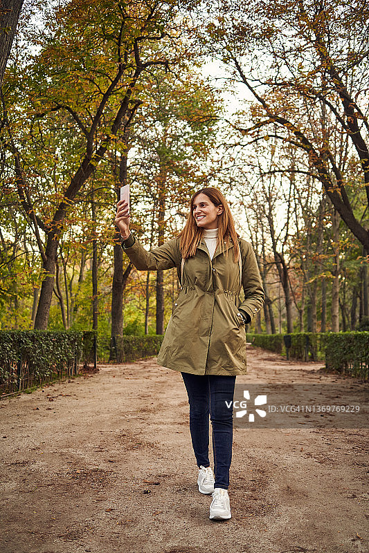 秋日，一名年轻女子在树木环绕的公园里用手机自拍。图片素材