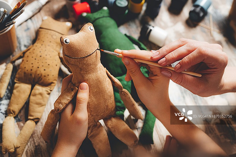 母亲和孩子的手着色手工玩具。和孩子们一起创造。制作老式玩具的过程。手工制作的青蛙玩偶。DIY制作的礼物。特写镜头图片素材