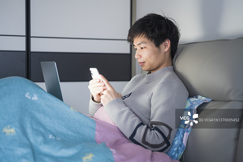 晚上，一名年轻的亚洲男性坐在床上用笔记本电脑工作图片素材