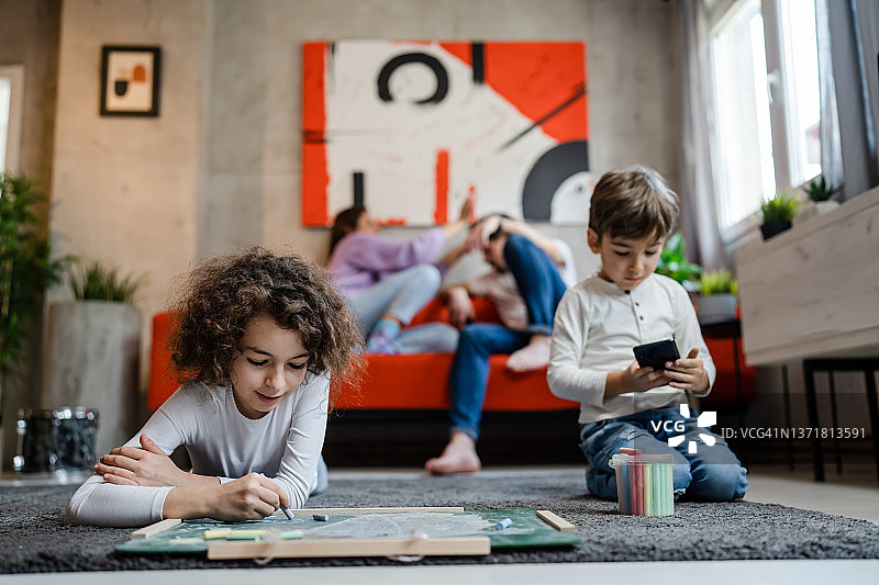小男孩和妹妹在家里的绿木板上画画，两个孩子的兄弟姐妹和父母躺在地板上用粉笔休闲教育真人的家庭观念图片素材