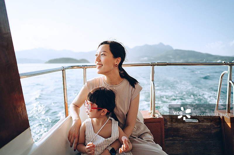 年轻的亚洲母亲和可爱的小女儿，戴着红色的太阳镜，在一个夏日的平静的海上享受游艇。眺望大海，放松心情。家庭生活方式。享受户外乐趣，乘船旅行和海上度假图片素材