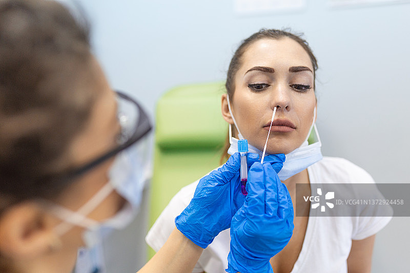 身穿防护服的医生正在对一名年轻女性进行鼻拭子测试。戴口罩的女性患者为了接受新冠病毒感染症(Covid - 19) pcr检测，正在接受鼻子样本采集。图片素材