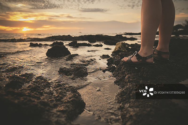 一个人在日落时穿着凉鞋站在岩石海滩上的特写图片素材