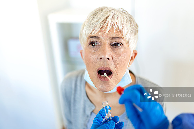 在临床环境中，一名穿着防护服的卫生保健工作者正在擦拭一名成年白人妇女的棉签，以确定她是否感染了冠状病毒或COVID-19。图片素材