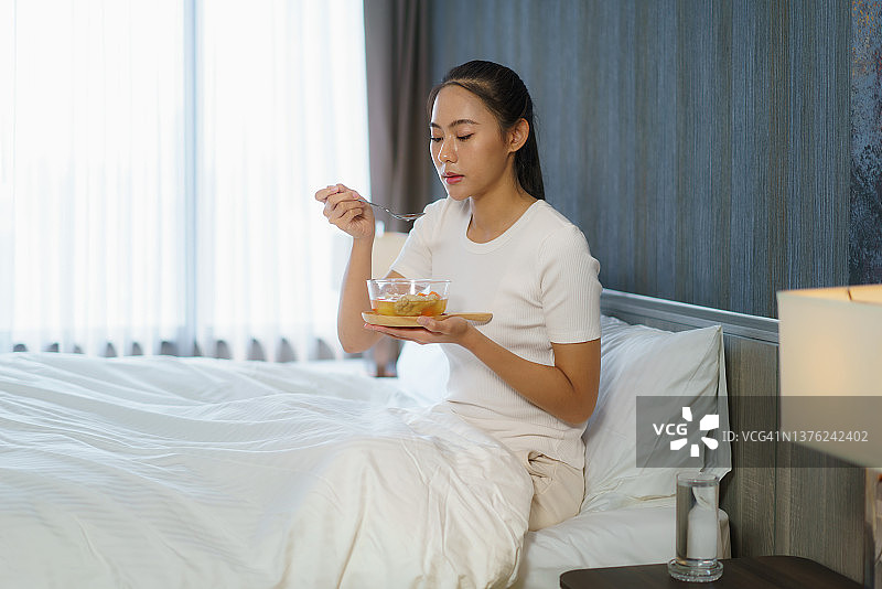 一位喉咙痛的亚洲妇女穿着睡衣在床上吃鸡汤来缓解喉咙痛。在家里的卧室里图片素材