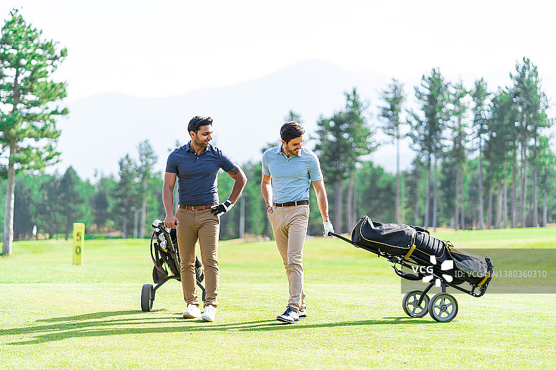 图为，在高尔夫球场上，拿着高尔夫球杆，拉着带轮子的高尔夫球袋，面带微笑的年轻高尔夫球手们。图片素材