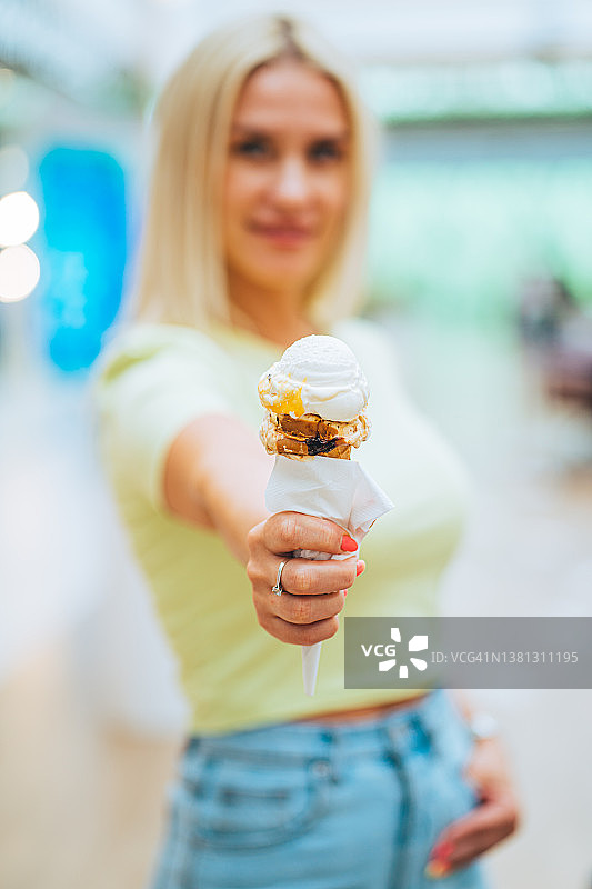 在一家购物中心，一个模糊的年轻金发女子用餐巾拿着一个黄白色的华夫饼蛋筒。美味可口的甜食。图片素材